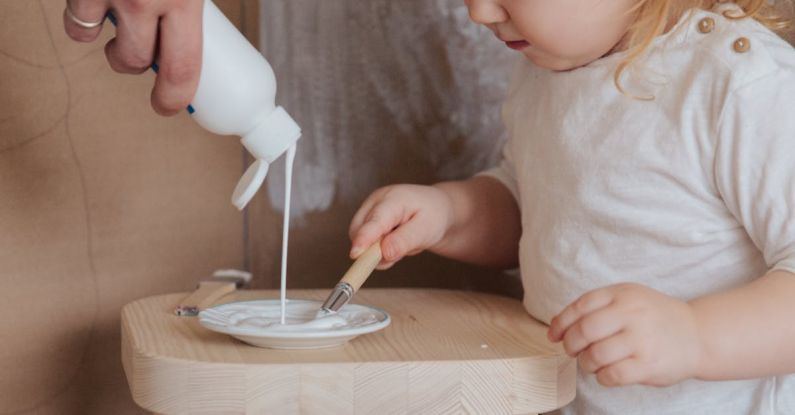 Game Flow - Crop faceless cute toddler girl holding brush for getting white paint flowing from bottle in parent hand to plate standing on wooden chair at home