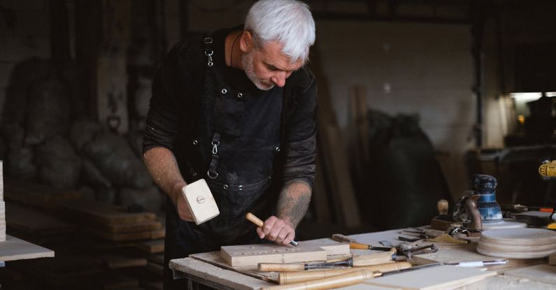Special Edition Board Game Releases - Serious male woodworker in apron using mallet and chisel to carve wooden board at table with abundance of instruments in carpentry