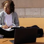 Online Multiplayer Platforms - Contemplative African American schoolkid with copybook and netbook sitting with crossed legs on wooden platform while looking away in sunlight