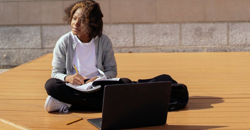 Online Multiplayer Platforms - Contemplative African American schoolkid with copybook and netbook sitting with crossed legs on wooden platform while looking away in sunlight